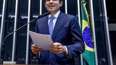 Quem é Hugo Motta discursando na Presidência da Câmara dos Deputados, segurando documentos diante da bandeira do Brasil.